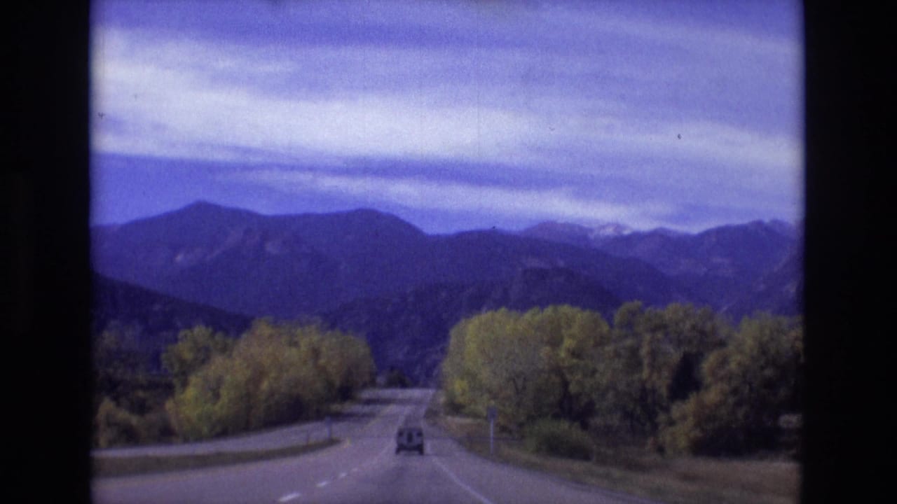 Two-lane road curving toward distant mountains with a car ahead and trees along the roadside.