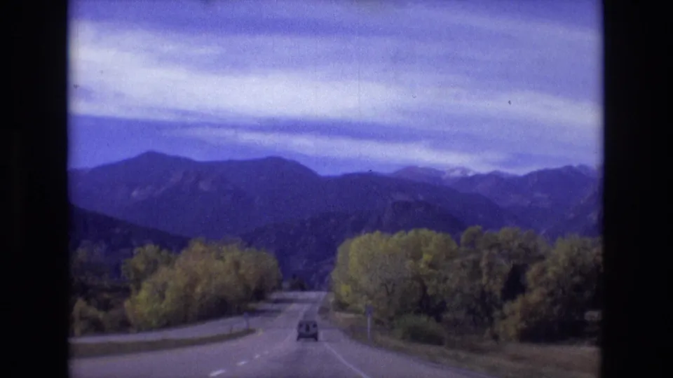 1974 Colorado Springs 8mm frame showing a moving-vehicle view along a two-lane road toward distant mountains under a cloudy sky.