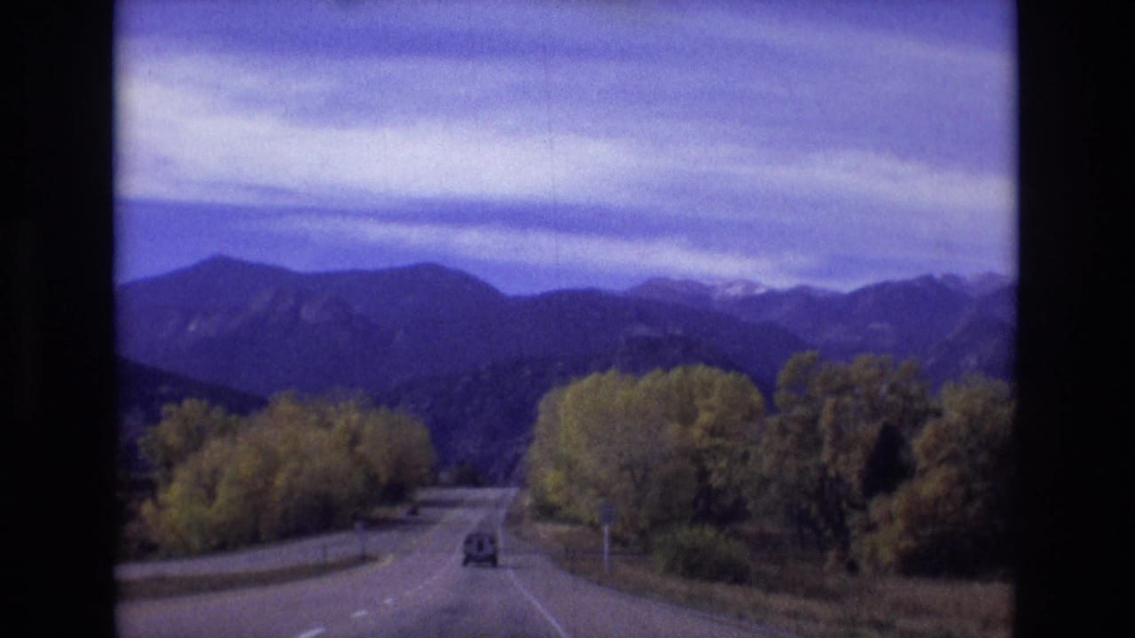 Vehicle-view road scene with a car ahead, mountain range in the distance, and trees beside the road.