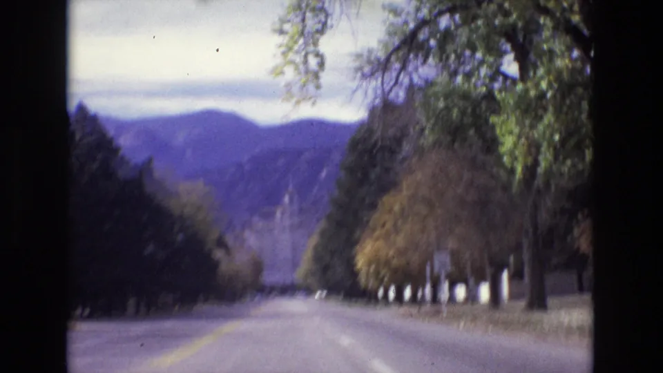 1974 Colorado Springs 8mm frame from a moving vehicle on a straight tree-lined road with mountains beyond and a building partly obscured by trees.