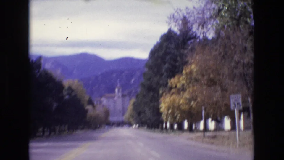 1974 Colorado Springs vehicle-view frame showing a straight road, tree-lined edges, distant mountains, a roadside sign, and a partly visible building at right.