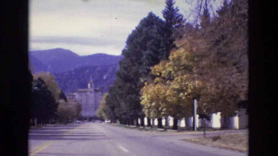 1974 Colorado Springs 8mm frame showing a quiet tree-lined boulevard, yellow foliage, a distant towered building, and mountains under an overcast sky.