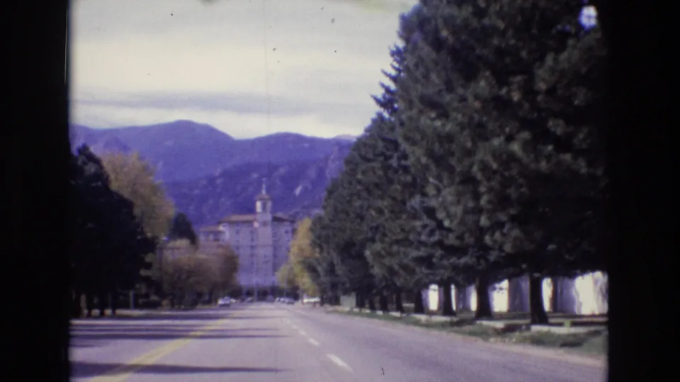 1974 Colorado Springs 8mm frame with a slightly closer roadside view of tall evergreens, a quiet paved road, a distant building, and purple-toned mountains.