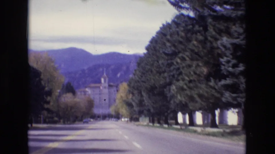1974 Colorado Springs 8mm frame showing the quiet boulevard continuing past tall evergreens and yellow trees toward a distant building beneath mountain ridges.