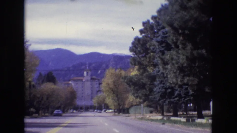 1974 Colorado Springs 8mm frame from a moving vehicle on a broad avenue, with roadside trees, a yellow lane line, distant traffic, mountains, and a towered building.