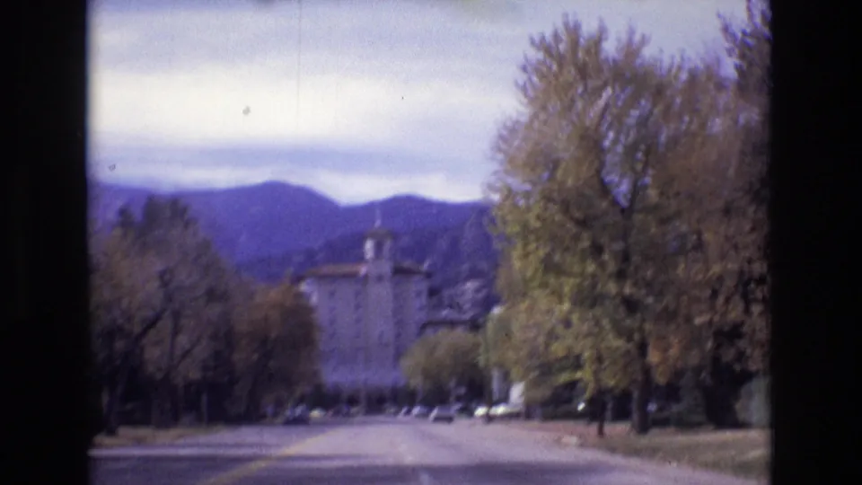 1974 Colorado Springs home-movie frame showing a street bordered by autumn-toned trees, distant cars, mountains, and a partially obscured towered building.
