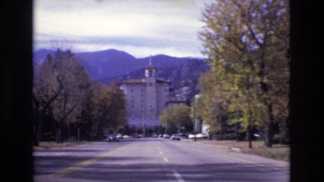 Wide road lined with trees leading toward a large hotel and mountains.