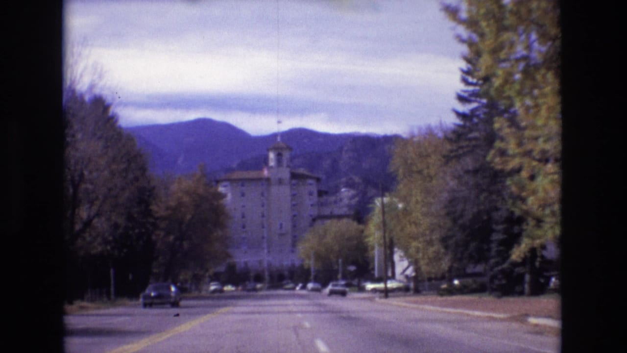 1974 Colorado Springs frame from a moving road view toward the Broadmoor Hotel, with autumn trees, parked or moving cars, and mountain ridges behind the building.