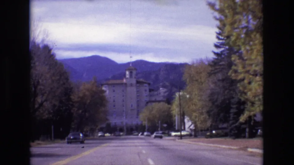 1974 Colorado Springs frame from a moving road view toward the Broadmoor Hotel, with autumn trees, parked or moving cars, and mountain ridges behind the building.