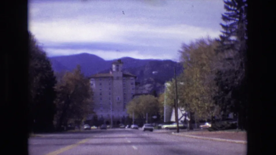 1974 Colorado Springs home-movie frame showing a vehicle-level view down the road toward the Broadmoor Hotel, with trees on both sides and mountains behind it.