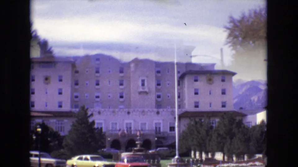 Closer 1974 Colorado Springs frame of the Broadmoor Hotel exterior, showing the multi-story facade, central tower, front drive area, cars, flagpole, and mountains beyond.