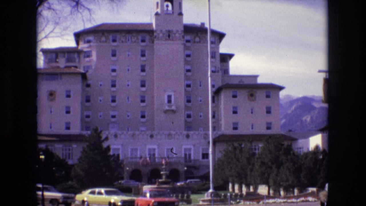 Large hotel facade with central tower, parked cars, trees, and mountains behind it.