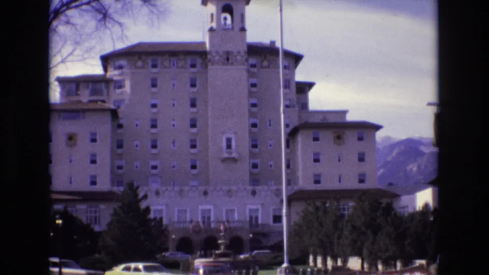 Final 1974 home-movie frame of the Broadmoor Hotel exterior in Colorado Springs, with the towered facade, parked cars, evergreen landscaping, flagpole, and mountains behind.