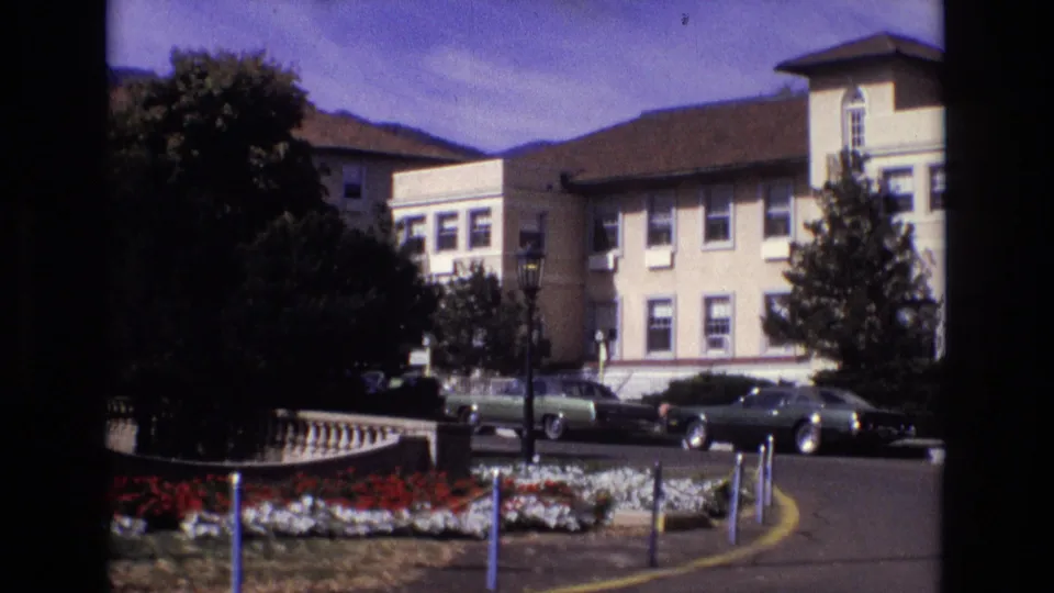 1974 Colorado Springs home-movie frame showing Broadmoor Hotel grounds with flower beds, a curved driveway, trees, adjacent hotel buildings, and parked period cars.