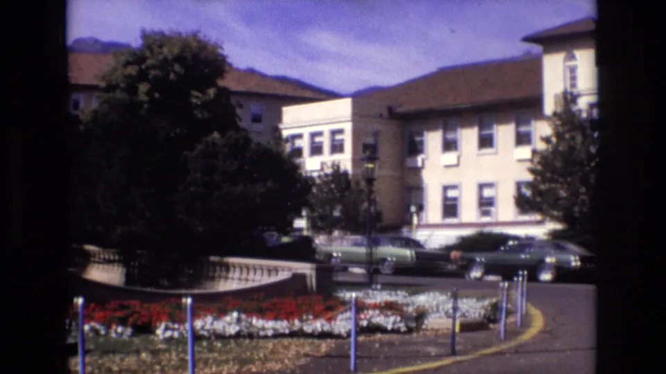 1974 Colorado Springs frame of the Broadmoor Hotel grounds, showing flower beds, a curved drive, trees, parked classic cars, and hotel buildings below the mountains.