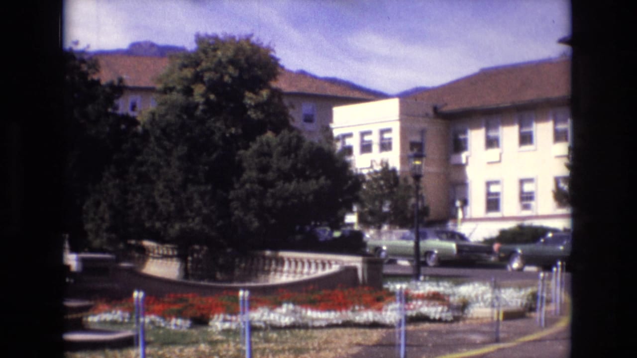 1974 Colorado Springs home-movie frame showing red and white flower beds, mature trees, parked cars, beige buildings, and distant mountains beyond a landscaped civic area.