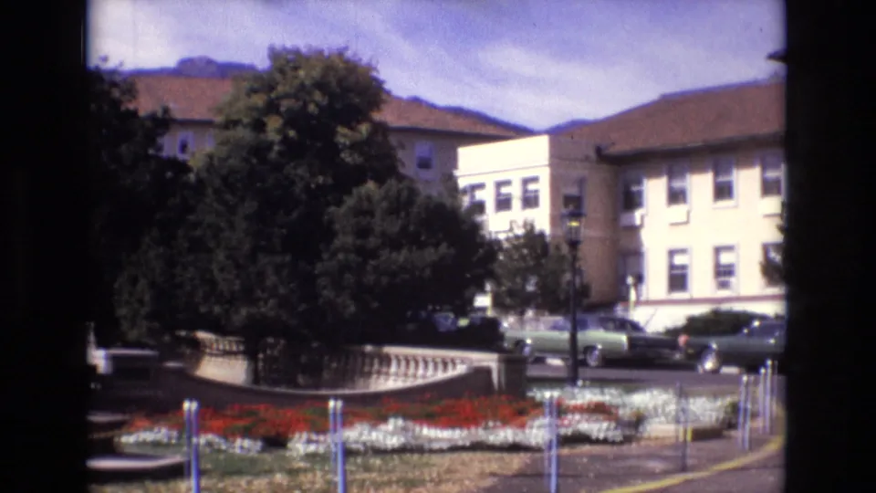 1974 Colorado Springs home-movie frame showing red and white flower beds, mature trees, parked cars, beige buildings, and distant mountains beyond a landscaped civic area.