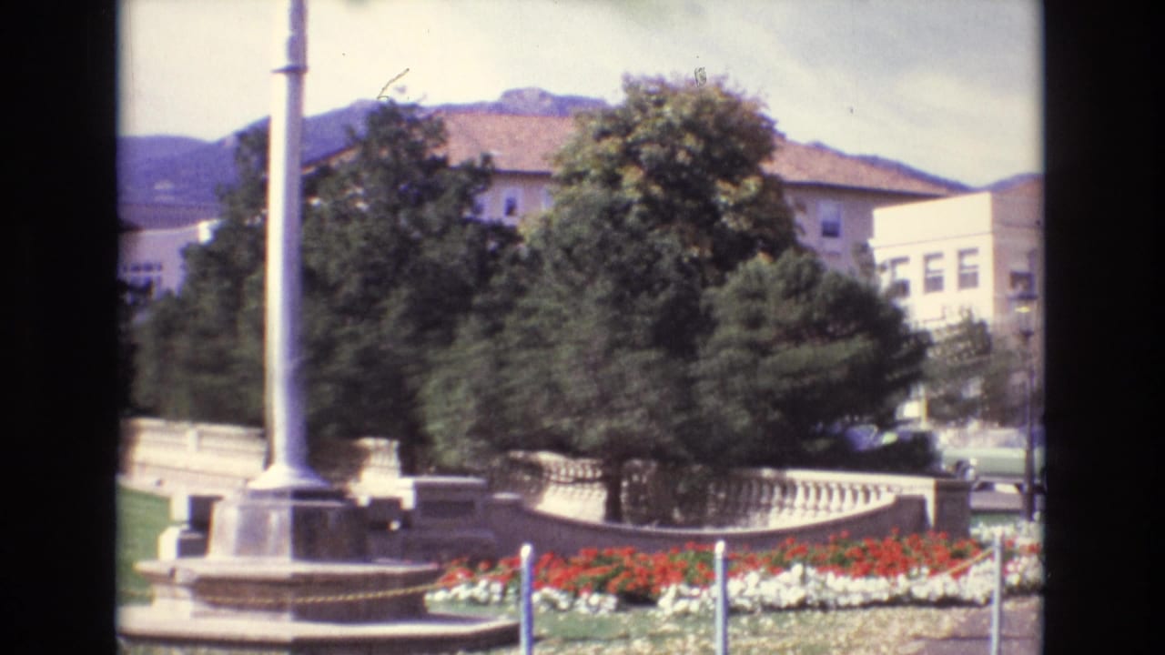 1974 Colorado Springs frame centered on a tall flagpole monument with flower beds, trees, parked cars, beige buildings, and mountain slopes behind it.
