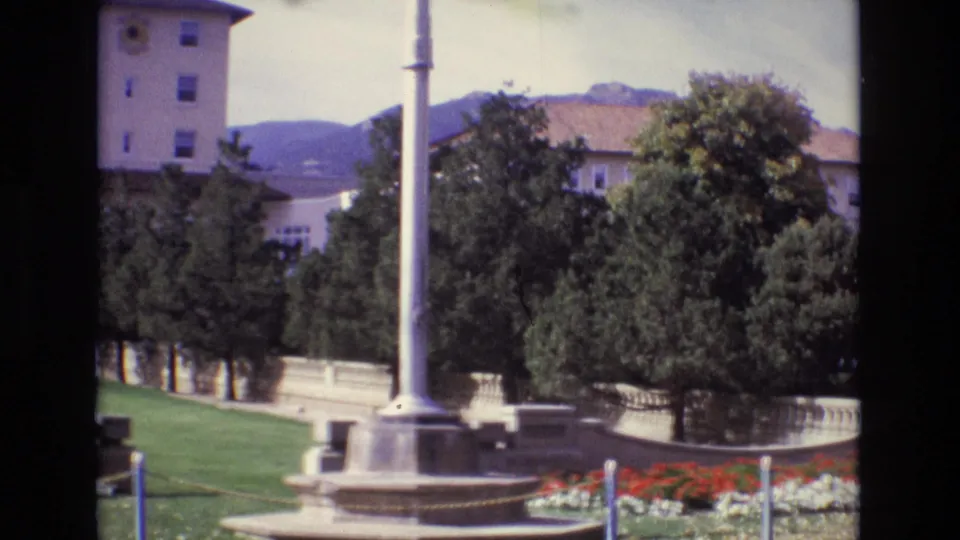1974 Colorado Springs home-movie frame showing the flagpole base, manicured lawn, red flowers, stone balustrade, trees, and a multi-story beige building near the mountains.