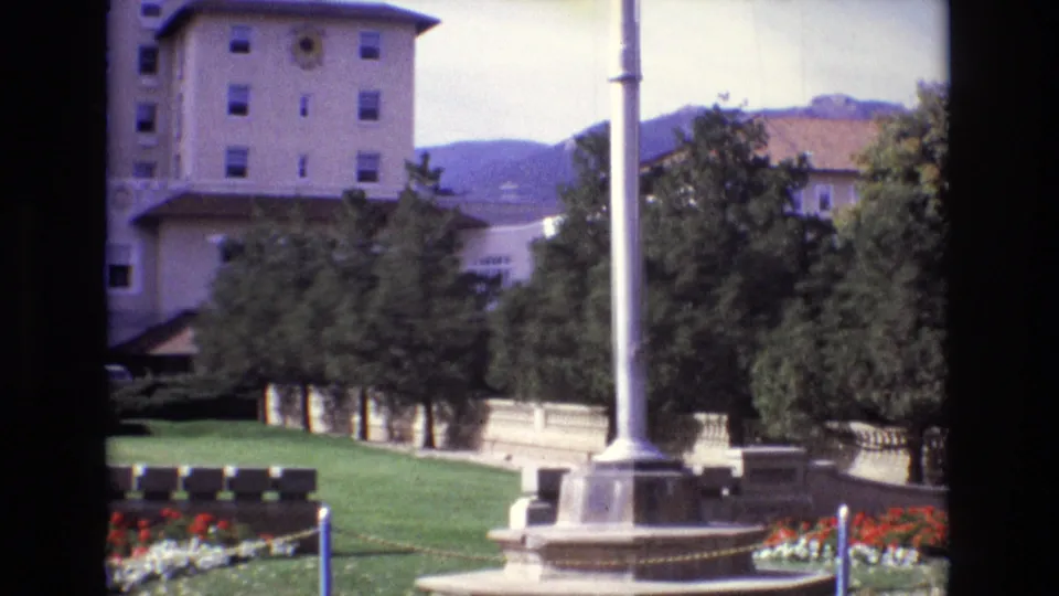1974 Colorado Springs frame with a flagpole monument in the foreground, benches on the lawn, red and white flower beds, trees, beige buildings, and distant mountains.