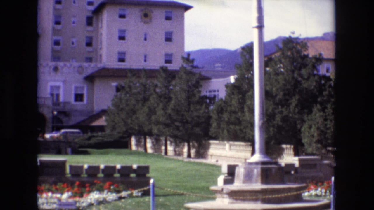 1974 Colorado Springs home-movie frame showing a grand hotel facade behind a landscaped courtyard with lawn, flower beds, trees, and a fountain base.