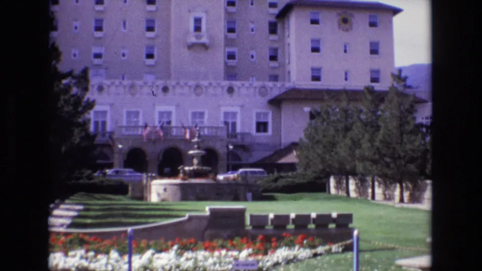 1974 Colorado Springs home-movie still showing a closer view of the hotel facade and fountain with benches, flower beds, lawn, trees, and parked cars.