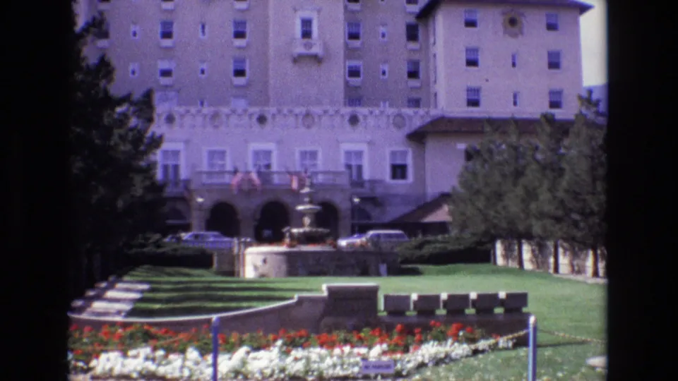 1974 Colorado Springs frame showing the hotel courtyard from a visitor viewpoint, with fountain, arched entrance, flower beds, lawn, trees, benches, and cars.
