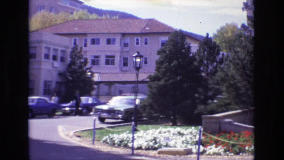 1974 Colorado Springs resort exterior with cars near the drive, flowerbeds and trees in the foreground, and small distant people near the hotel grounds.