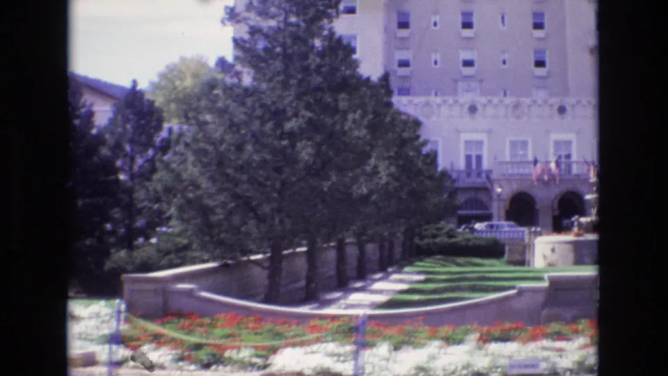 1974 Colorado Springs home-movie frame showing the hotel lawn, red and white flower beds, shaded path, stone wall, trees, and a large arched facade.