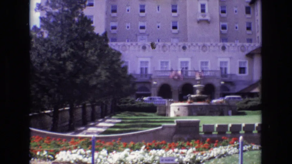 1974 Colorado Springs hotel grounds with a formal lawn, flower beds, arched entrance facade, parked cars, lampposts, and small distant visitors near the building.