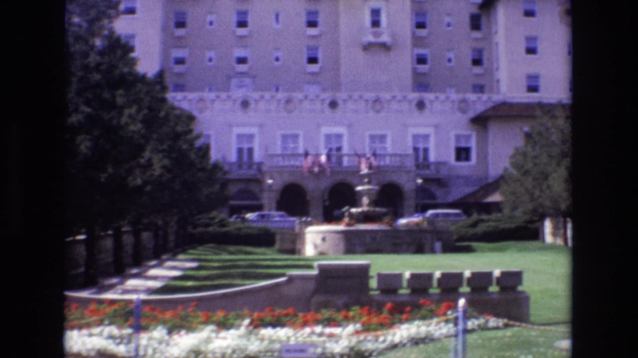 1974 8mm view of a large Colorado Springs hotel exterior with American flags above an arched entrance, a fountain, manicured lawns, flower beds, trees, and parked cars.