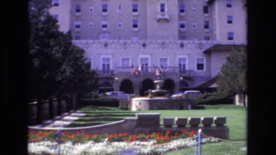 1974 8mm view of a large Colorado Springs hotel exterior with American flags above an arched entrance, a fountain, manicured lawns, flower beds, trees, and parked cars.