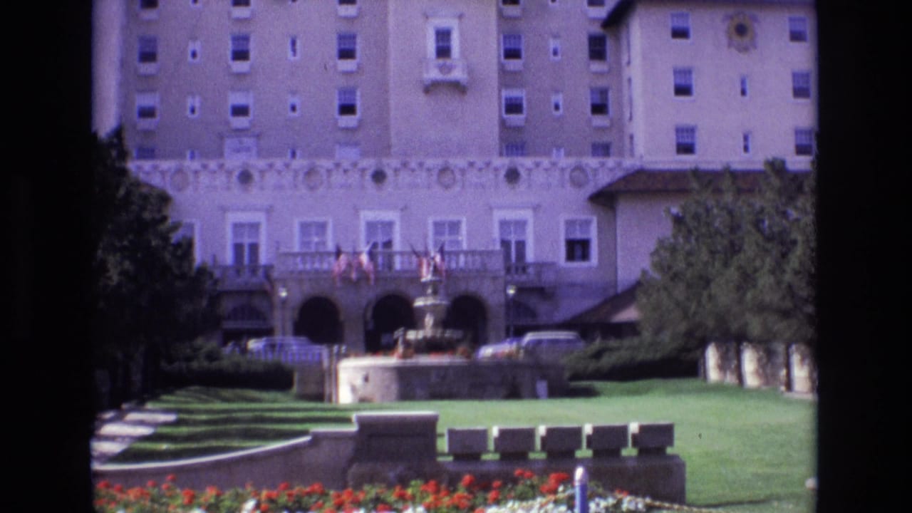 Hotel exterior seen across lawns and flower beds, with flags, fountain, trees, and cars near the entrance.