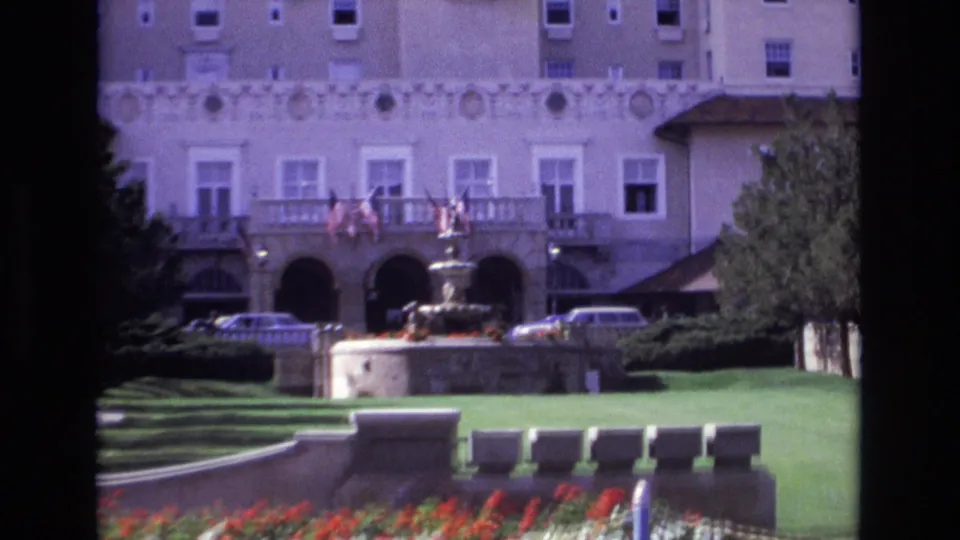 Closer 1974 8mm frame of the hotel entrance in Colorado Springs, with balcony flags, a fountain in front, formal lawn, red and white flower beds, trees, and cars along the drive.