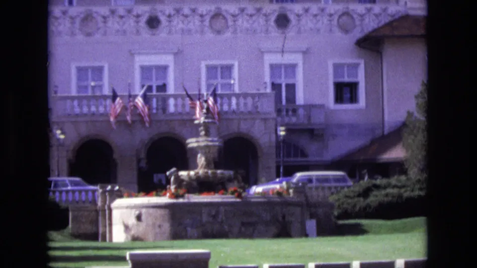 1974 Colorado Springs hotel exterior with arched doorways, balcony flags, a central fountain, clipped lawn, trees, and a parked station wagon or car near the entrance.