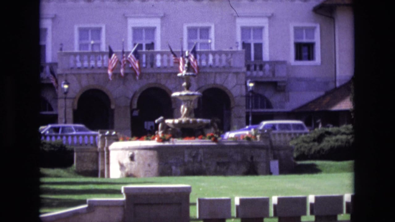 1974 Colorado Springs home-movie frame showing a formal pale building with arched entryways, balcony flags, a central fountain, green lawn, and parked cars.