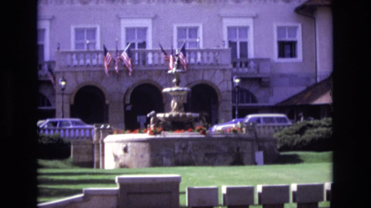 1974 Colorado Springs frame with the fountain centered before a large arched building, American flags on the balcony, lawn in the foreground, and parked 1970s cars nearby.