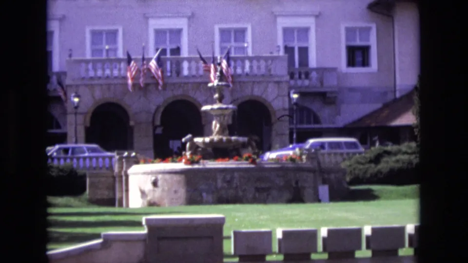 1974 Colorado Springs home-movie frame holding on the fountain and formal building entrance, with flags above the arches, a trimmed lawn, flower planting, and cars at the curb.