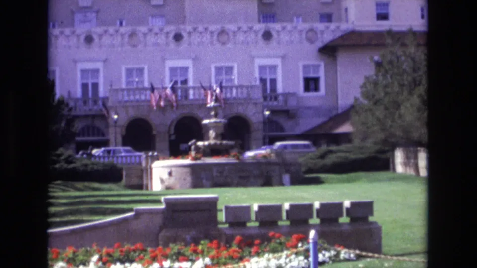 Wider 1974 Colorado Springs frame revealing more of the landscaped grounds, with red and white flowers, low stone walls, a central fountain, balcony flags, parked cars, and the formal building facade.