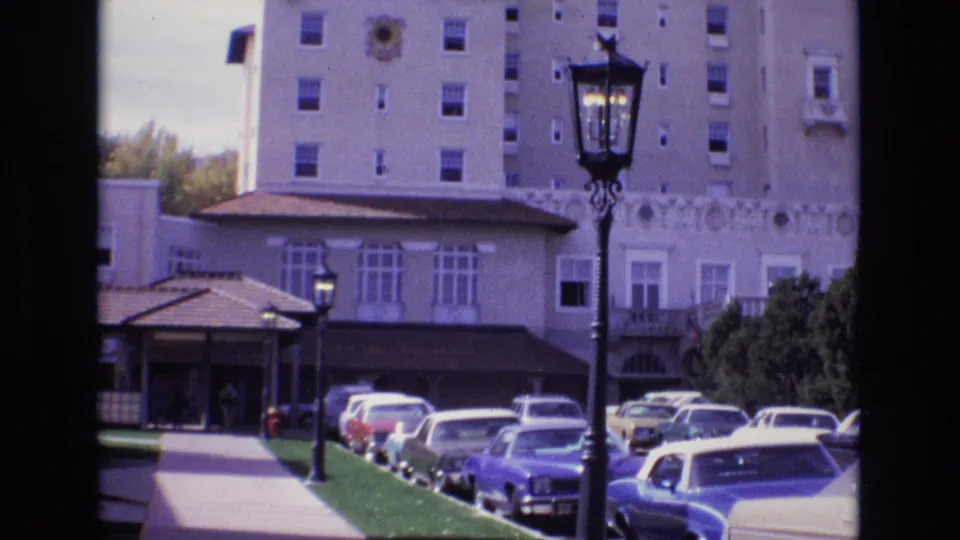 Closer 1974 8mm frame of the Broadmoor Hotel entrance area with parked cars along the driveway, a black lamppost, sidewalk, shrubs, and the upper hotel facade.