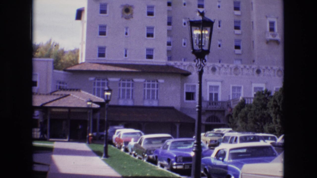 1974 Colorado Springs home-movie view of a tall tan hotel or large lodging building with a covered entrance, parked cars along the driveway, green lawn, and a wrought-iron style lamp post in front.