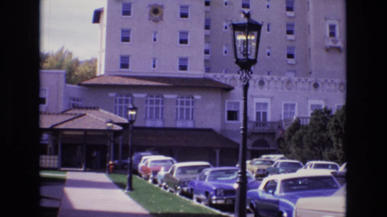 1974 Colorado Springs exterior shot showing a tan multi-story building, a covered arrival area, a row of classic cars, sidewalk, lawn, shrubs, and a prominent foreground lamp post.