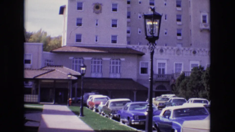 1974 Colorado Springs 8mm frame looking toward a covered hotel-style entrance with parked cars, a sidewalk and lawn strip, shrubs, and the upper stories of a tan building behind it.