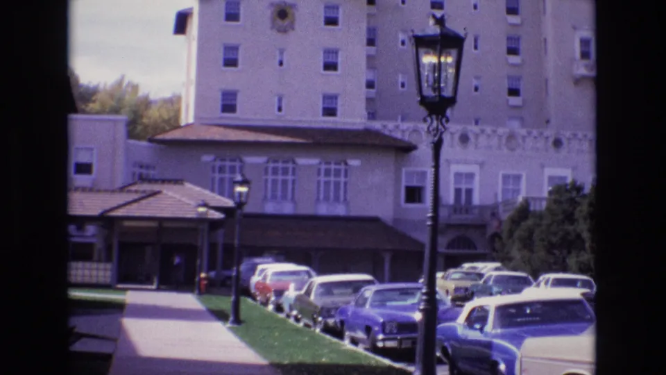 1974 Colorado Springs home-movie frame of a tall tan building with decorative facade details, a covered driveway entrance, parked automobiles, shrubs, lawn, and a dark foreground streetlight.
