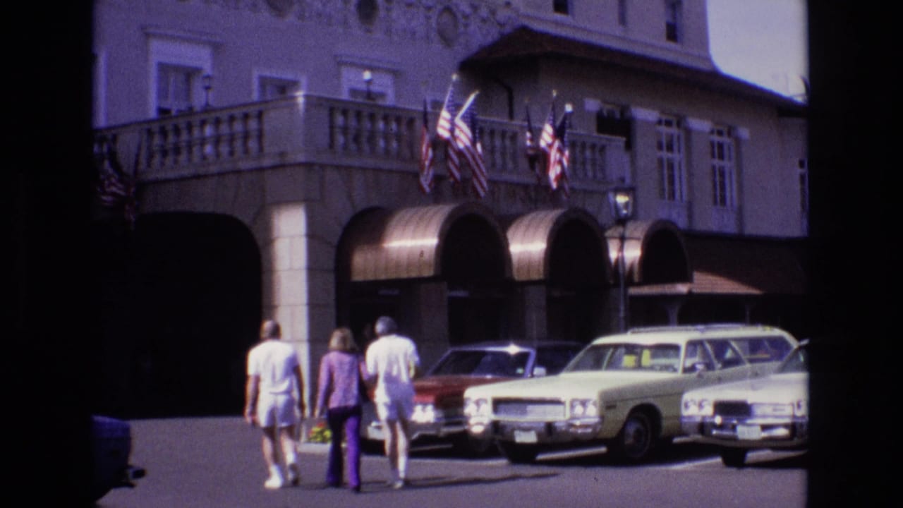 1974 Colorado Springs home-movie frame showing three adults approaching a large hotel facade with arched awnings, American flags, a yellow station wagon, and a red sedan in the parking area.