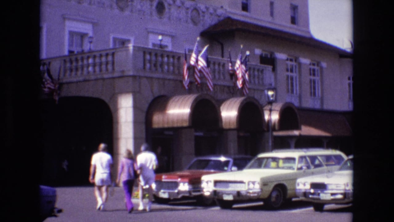 1974 Colorado Springs frame with three adults crossing the lot toward a large hotel entrance, with American flags above arched awnings and several period cars parked in front.