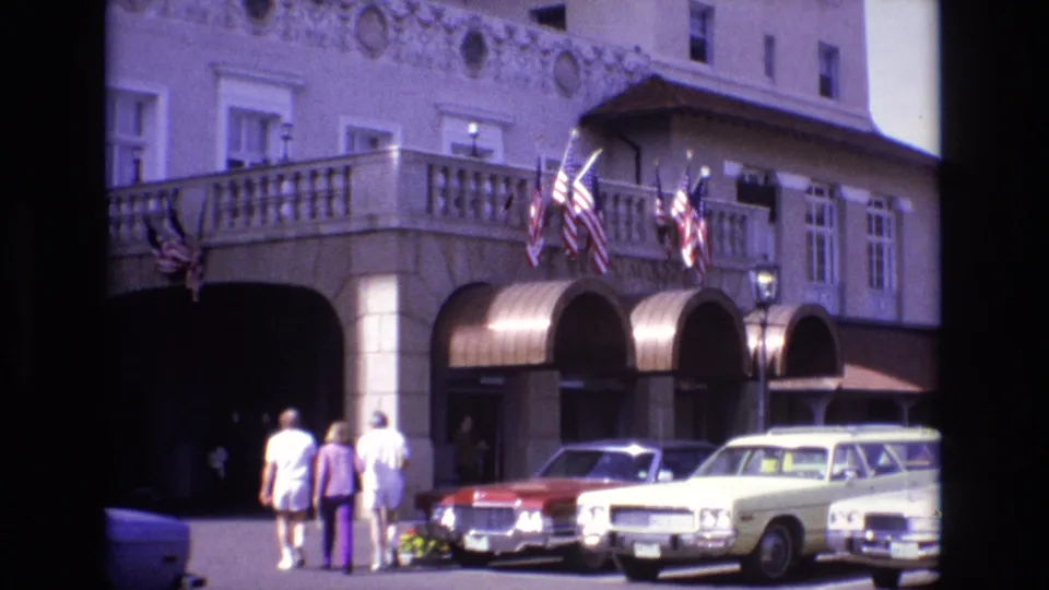 1974 Colorado Springs home-movie still showing three visitors near the shaded hotel entry, with American flags, arched awnings, flower planters, and 1970s automobiles in view.