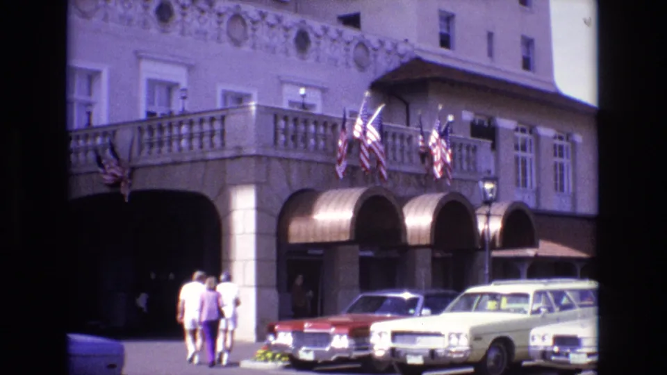 1974 Colorado Springs frame showing adults moving toward the arched hotel entrance, with American flags above the awnings and vintage cars parked along the front of the building.