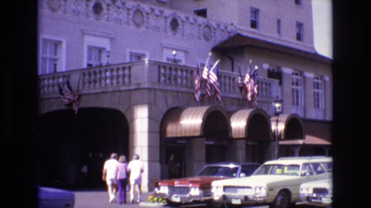 In 1974 Colorado Springs, three adults walk toward a grand arched entrance with American flags above a red sedan and cream-colored station wagon parked at the curb.