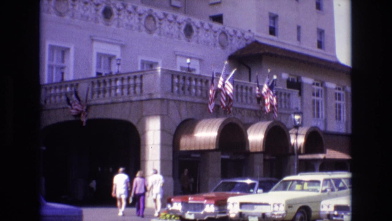 A 1974 Colorado Springs street view shows three pedestrians nearing a shaded arched building entrance while American flags hang above parked mid-century cars.
